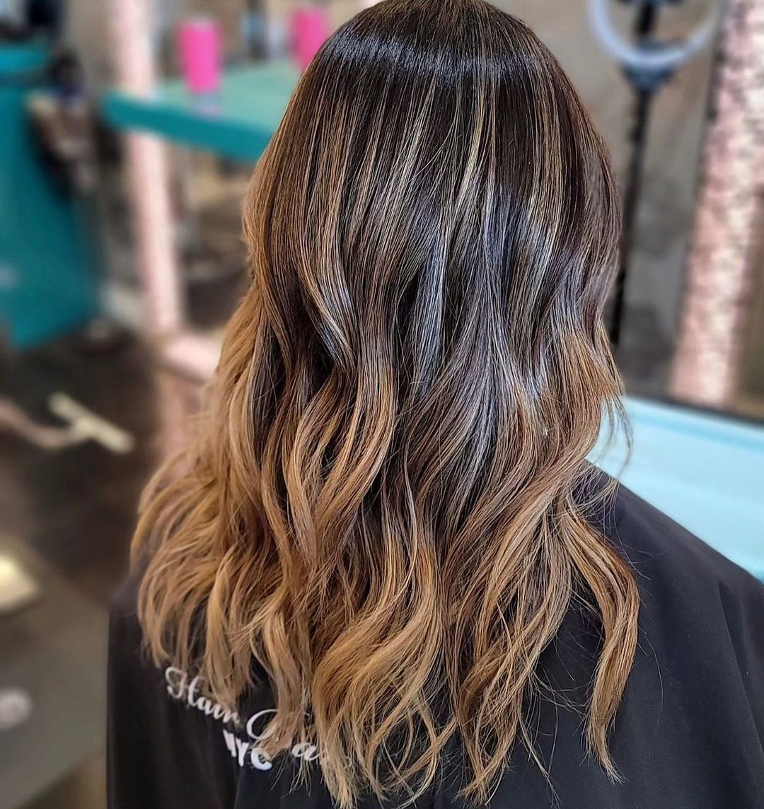 A person with long, wavy hair faces away from the camera. The hair is dark brown at the roots and transitions smoothly to lighter, caramel blonde at the ends, showing a balayage effect. They wear a black salon cape in a bright, modern salon setting.