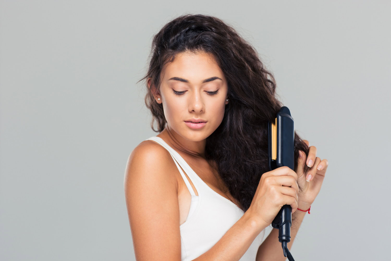 A woman with long, wavy dark hair and light skin uses a black hair straightener. She wears a white tank top, looking down with a neutral expression, against a plain light gray background.