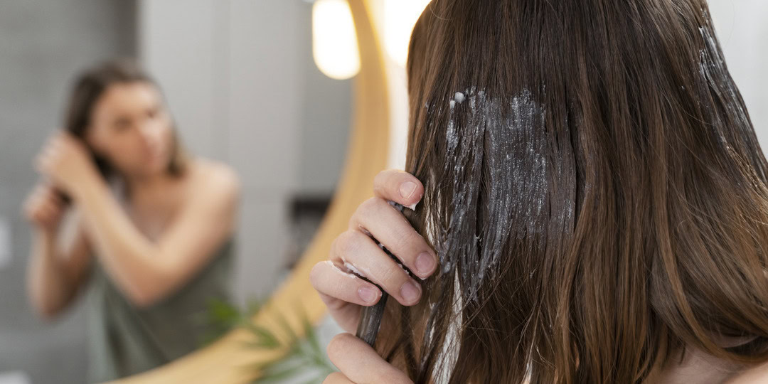 A person with long brown hair applies a white hair treatment or mask with their hand, partially covering their face. They stand in front of a mirror, where their blurred reflection is visible, in a bathroom setting with soft lighting.