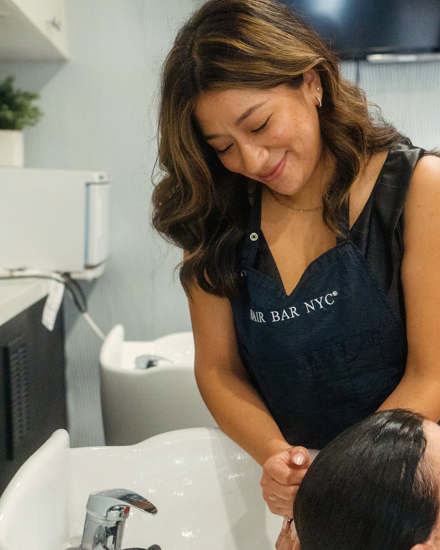 A hairstylist with wavy brown hair, wearing a “Hair Bar NYC” apron, smiles while washing a client’s hair at a salon sink. The background shows another sink, white cabinets, and a small green plant on a shelf.
