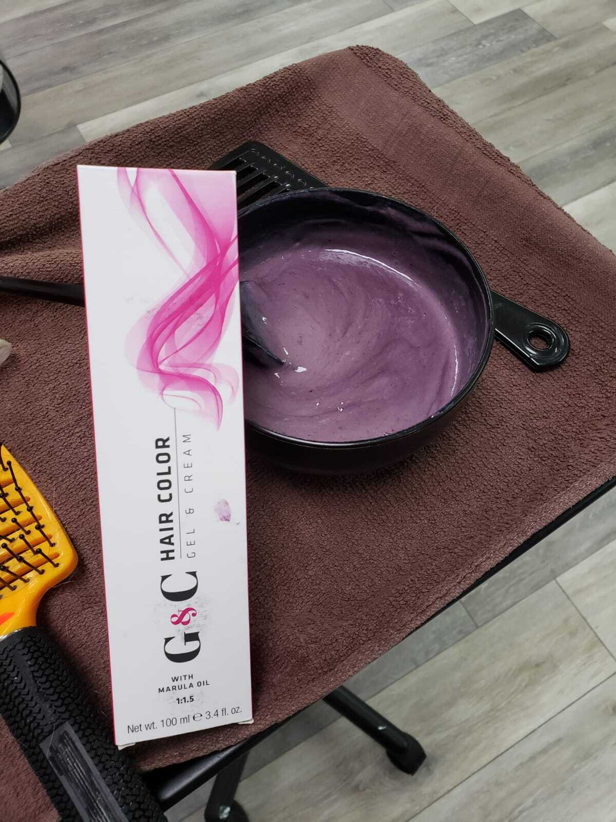 A black bowl with creamy purple hair dye sits on a brown towel, next to a box labeled G&G Hair Color with Marula Oil. A brush and yellow hair comb are nearby, all placed on a light wood-textured floor.