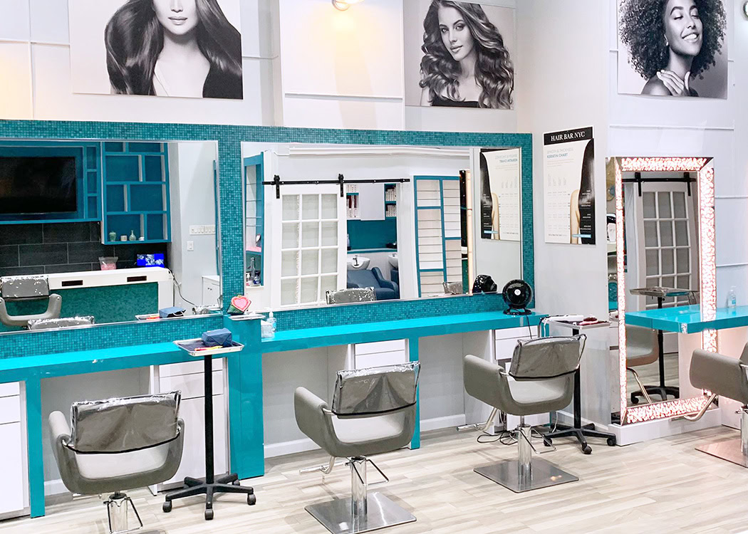 A modern hair salon features teal and white decor, four gray styling chairs facing large mirrors, and light wood floors. Posters of women with styled hair hang above. Tools, hair products, and a lit vanity mirror are visible on the counters.
