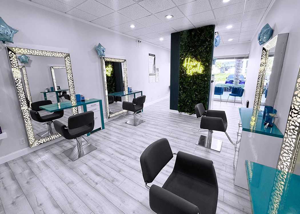 Modern hair salon with white walls and light wood floors. Three black styling chairs face ornate mirrors with intricate frames and teal countertops. A green plant wall with a “Hair Goals” neon sign is at the back, near large windows and more blue seating.