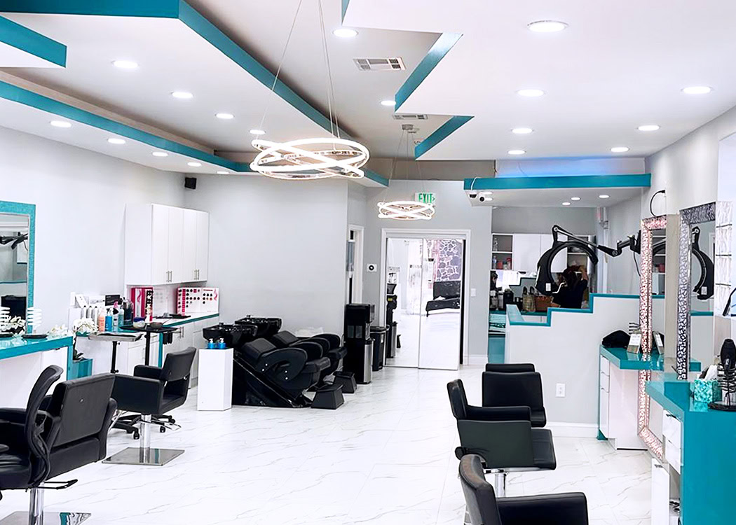 Modern hair salon interior with black styling chairs, white walls, turquoise accents, mirrors, and circular hanging lights. Stations are neatly arranged along the walls. The floor is white with a marble pattern, and large windows allow natural light in the back.