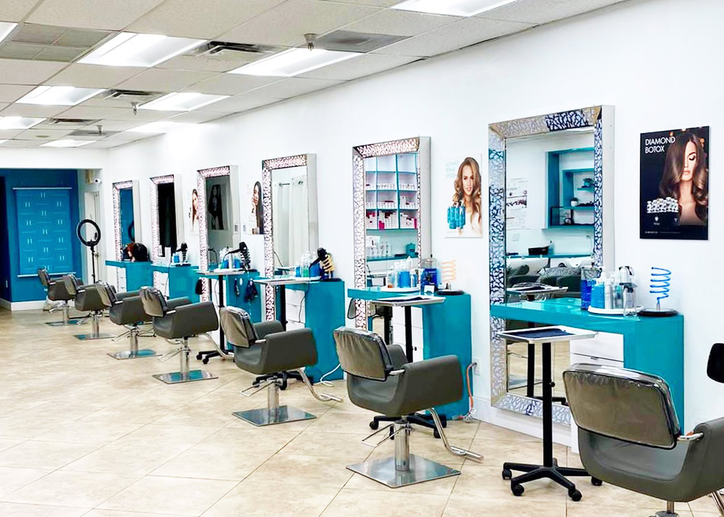 A modern hair salon with turquoise styling stations, large mirrors, and black chairs. Each station has hair products and tools. The salon has a tiled floor, white walls, and posters of models with styled hair. The space looks clean, organized, and well-lit.