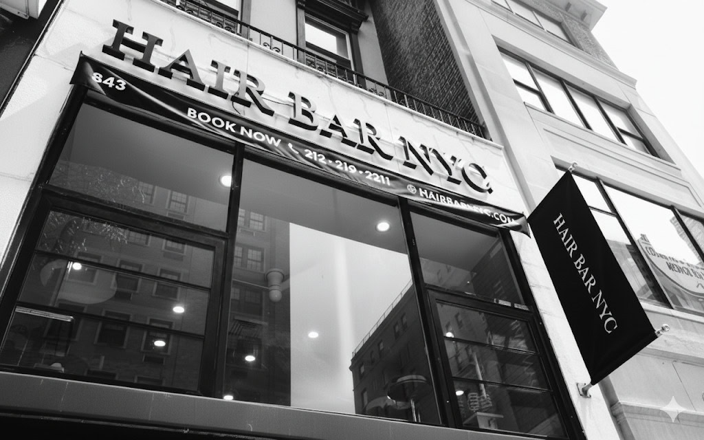 Black and white photo of the exterior of Hair Bar NYC, a hair salon. Large windows reflect surrounding buildings. The salon’s name and phone number are displayed on a sign above the entrance. A black side banner also reads “Hair Bar NYC.”.