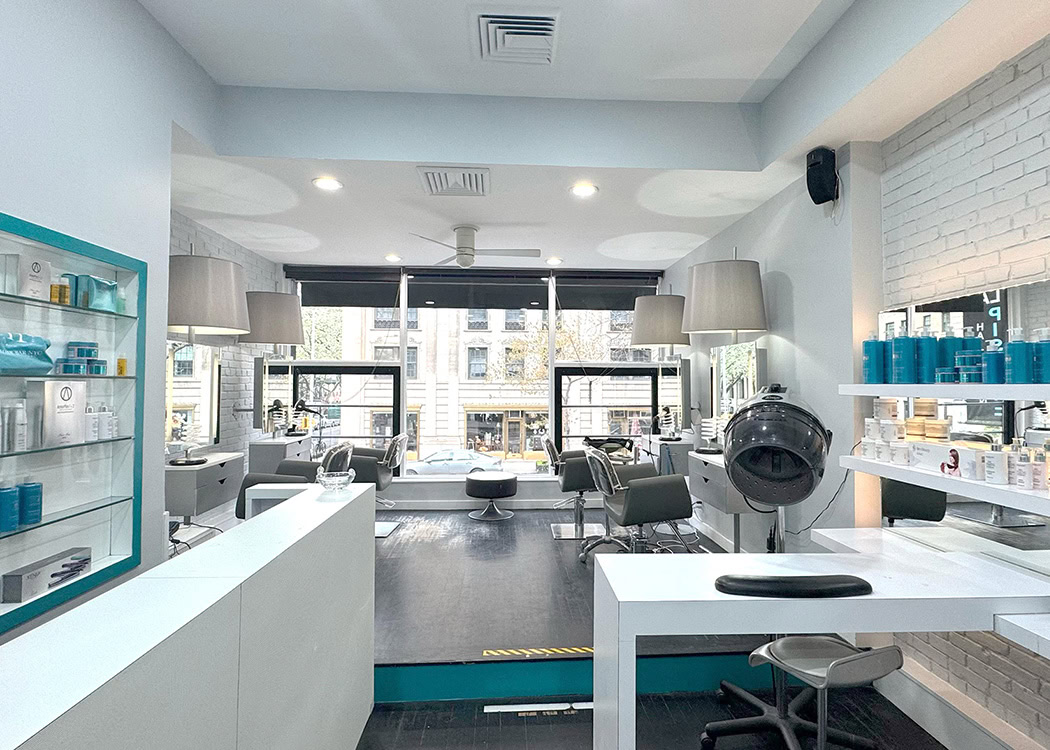 A modern, bright hair salon with large windows, white walls, and sleek decor. There are styling chairs, product shelves with bottles, a hair dryer station, and large mirrors. Outside, city buildings and trees are visible through the windows.
