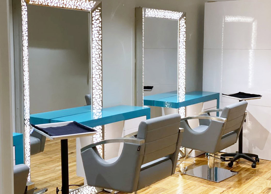 A modern hair salon interior with two styling stations. Each station has a turquoise countertop, a large rectangular mirror framed with bright, decorative lights, and a grey salon chair. The floor is wooden, and the walls are light-colored and glossy.