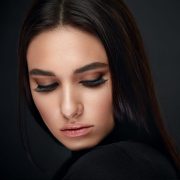 A woman with long dark hair and smooth skin looks down, eyes closed and bold brows framing her dramatic black eyeliner. Her lush lashes, styled with the 3D Faux Mink Eyelash Kit, stand out against her black top and the moody background.