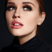 A woman with flawless skin, bold brows, glossy pink lips, and brown hair pulled back gazes upward. She wears a black turtleneck as soft light highlights her 3D Faux Mink Eyelash Kit lashes against a dark background.