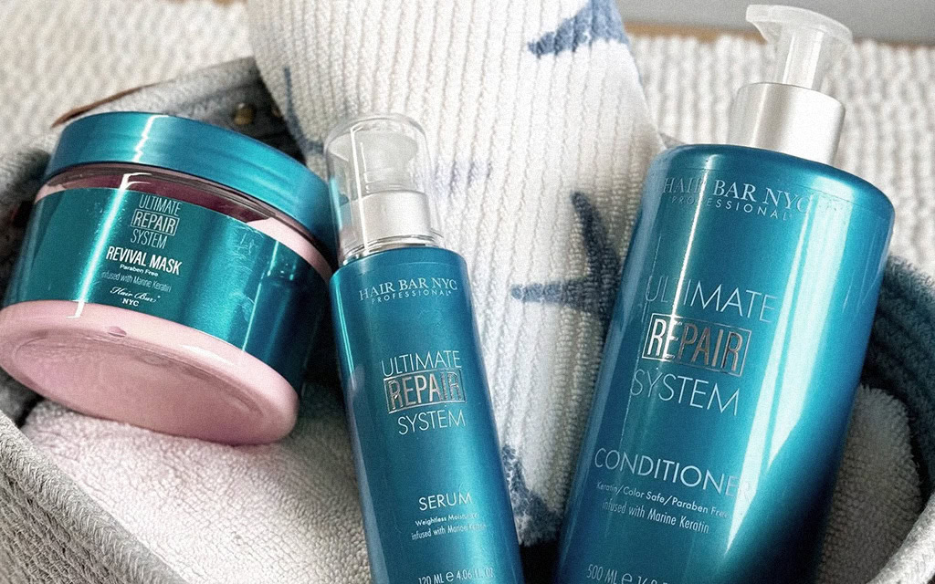 Three turquoise Hair Bar NYC “Ultimate Repair System” products—Revival Mask (jar), Serum (pump bottle), and Conditioner (large pump bottle)—are arranged on a white textured towel with blue designs, inside a light gray basket. Three turquoise Hair Bar NYC “Ultimate Repair System” products—Revival Mask (jar), Serum (pump bottle), and Conditioner (large pump bottle)—are arranged on a white textured towel with blue designs, inside a light gray basket.