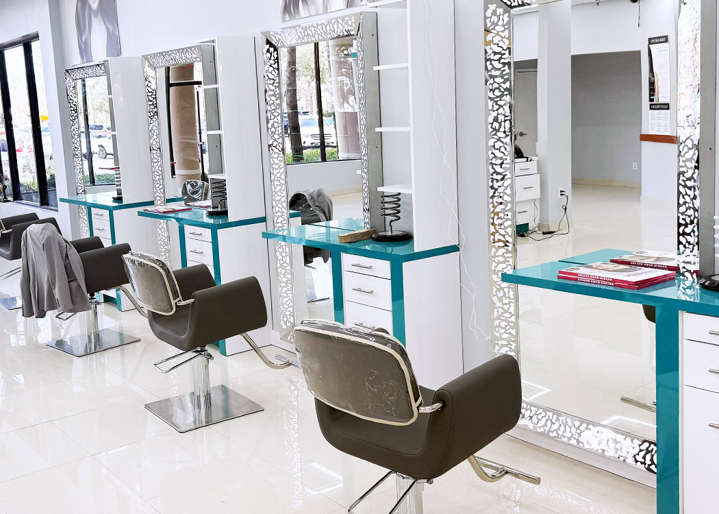 A modern, brightly lit hair salon with large mirrors framed by metallic patterns, teal countertops, and brown swivel chairs. Each station has a hairdresser’s chair, shelves, drawers, and hairdressing tools, with jackets draped over some chairs.