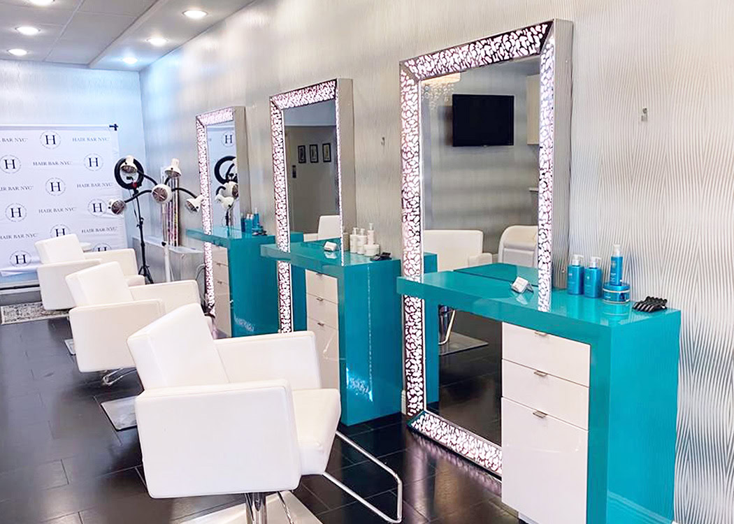 A modern hair salon with three turquoise styling stations, each with a large mirror framed by lights. White salon chairs are positioned at each station. Shelves display hair products, and the floor is dark with a glossy finish. The walls are light-colored.