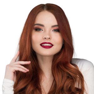 A woman with long, wavy auburn hair and fair skin smiles softly, showcasing her fresh Single Process Color (Root Touch-up). She wears burgundy lipstick, rosy eyeshadow, and a white top, posing with one hand raised near her face against a white background.