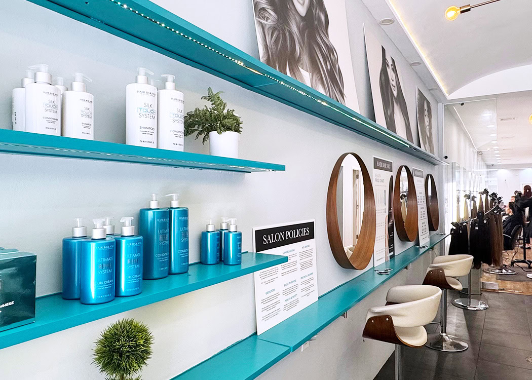 Modern hair salon interior with turquoise shelves displaying hair products and small plants, round mirrors above white and brown salon chairs, and black-and-white portraits of women with styled hair on the wall. Salon policies sign is visible on the counter.