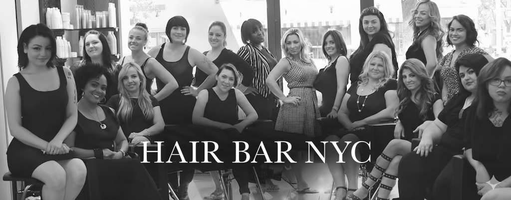 A group of seventeen women, mostly in black outfits, pose together inside a hair salon. They are smiling, with salon shelves in the background. Bold text across the bottom center reads “HAIR BAR NYC.” The image is in black and white.