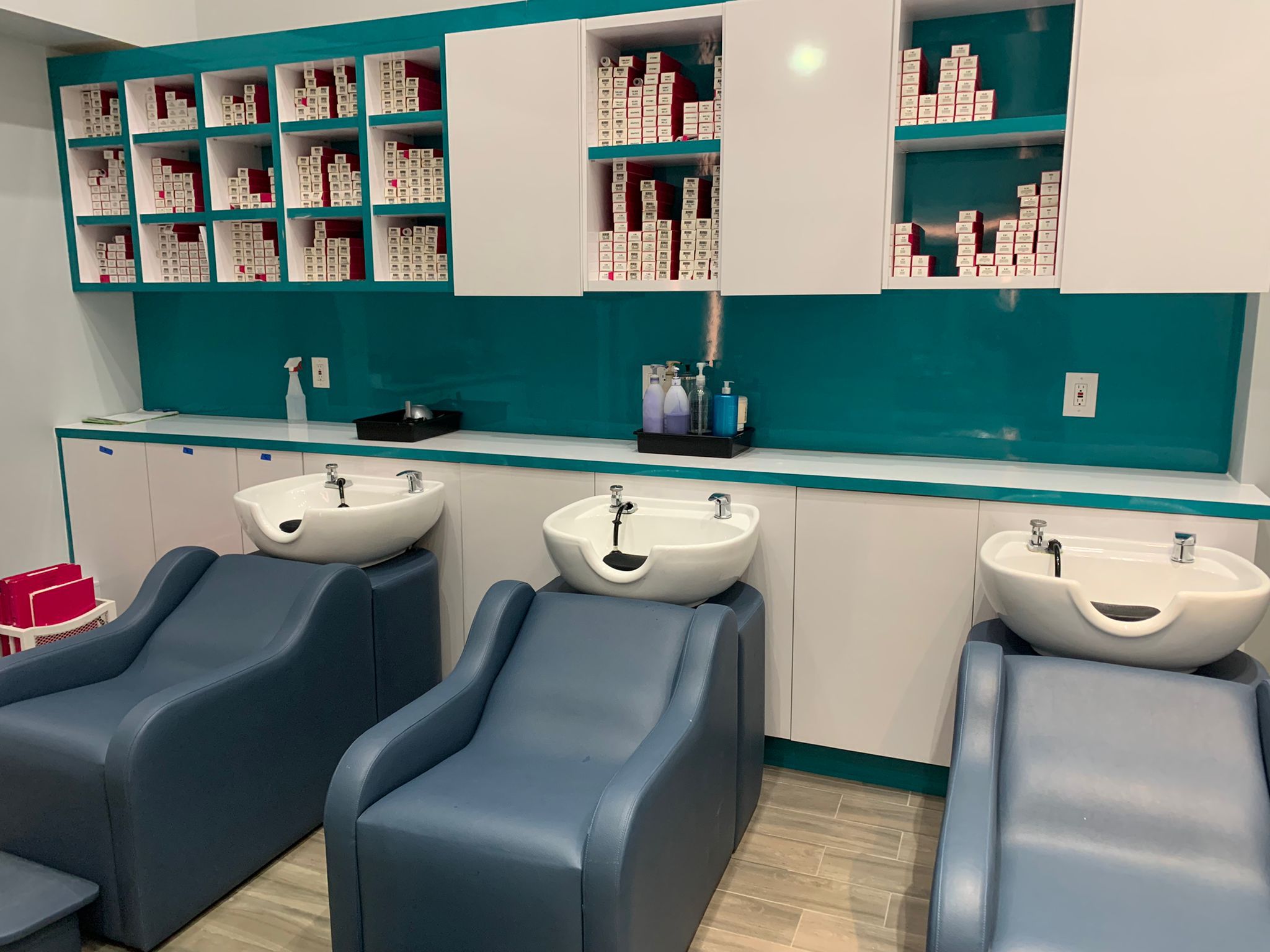 Three modern salon wash stations with blue chairs and white sinks are in front of a teal wall. Shelves above hold organized rows of hair dye boxes, mostly red and white. Bottles and supplies sit on the counter beneath the shelves. The floor is light wood.