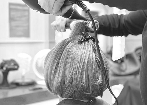 A person blow-dries the hair of another person in a salon. The focus is on the back of the client’s short, straight hair, sectioned and clipped. The stylist’s hands hold a blow dryer and lift a section of hair. The background shows salon equipment out of focus.