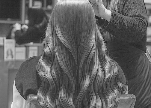A person with long, smooth, wavy hair sits in a salon chair, viewed from behind. A stylist, standing to the right, styles the hair, which appears glossy and well-groomed. Salon mirrors and shelves are visible in the background. The image is in black and white.
