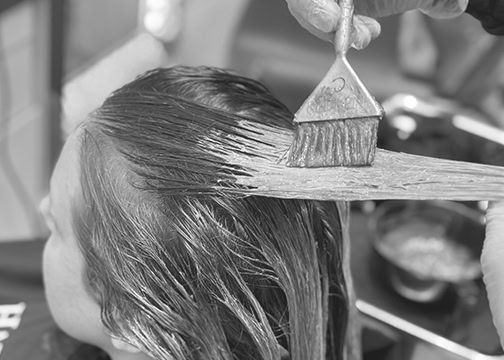 A person is having hair dye applied to their hair with a brush by someone wearing gloves. The hair appears wet and parted as the dye is spread. The scene is in a salon, with dye bowls and tools visible in the blurred background. The image is in black and white.