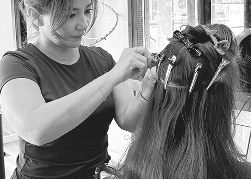 A hairstylist attaches hair extensions to a client’s long hair using clips. The client sits facing away, hair sectioned and clipped. The stylist focuses on her work, wearing a short-sleeved shirt in a bright, well-lit salon.