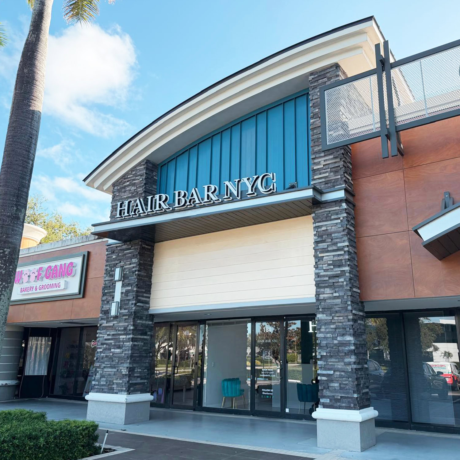 Storefront of “HAIR BAR NYC” in a modern strip mall, featuring tall stone pillars, large glass windows, and blue accents. Palm tree leaves and a pet grooming business sign are visible to the left under a bright, partly cloudy sky.
