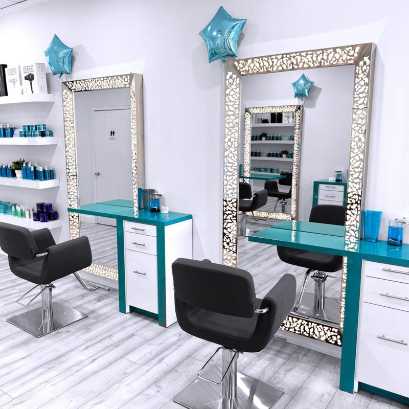 A modern hair salon with two black styling chairs facing large, ornate, backlit mirrors framed with cut-out patterns. Each station has teal and white cabinets. Blue star balloons decorate the mirrors. Shelves with hair products and a restroom door are visible.