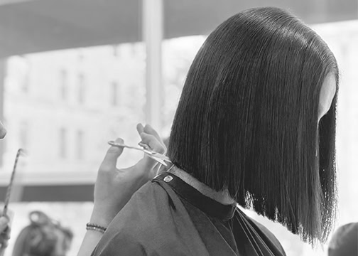 A person with straight, shoulder-length hair sits in a salon as a stylist uses scissors to trim the blunt ends of their sleek, asymmetrical bob haircut. The background shows large windows with blurred city buildings. The image is in black and white.
