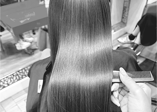 A person with long, straight, smooth hair sits in a salon chair. Another person’s hand holds a comb near the ends of the hair, preparing to style or cut it. The shiny hair reflects light, and other clients and salon equipment are visible in the background.