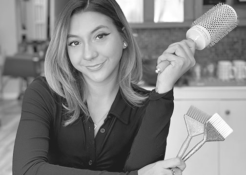 A young woman with shoulder-length hair, wearing a dark long-sleeve top, smiles at the camera. She holds a round hairbrush in one hand and two hair dye brushes in the other. The background shows a blurred indoor setting with shelves and kitchen items.