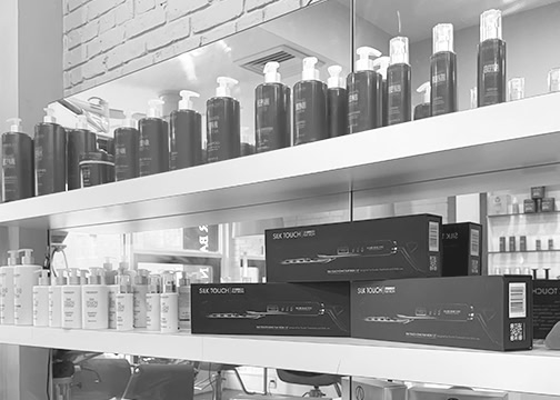 Black and white photo of salon shelves with rows of bottles, mostly pumps, and boxed hair styling tools. Reflections in the mirror show more products and salon chairs. The scene is neat and organized, with a modern, professional vibe.